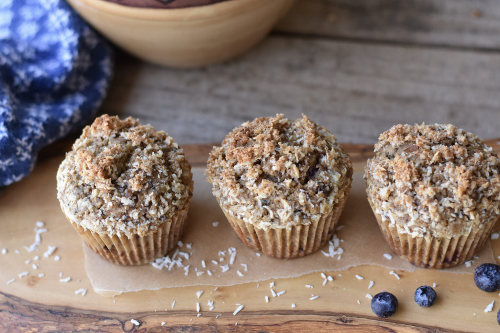 Three Grain free blueberry collagen muffins on a cutting board with blueberries beside them.