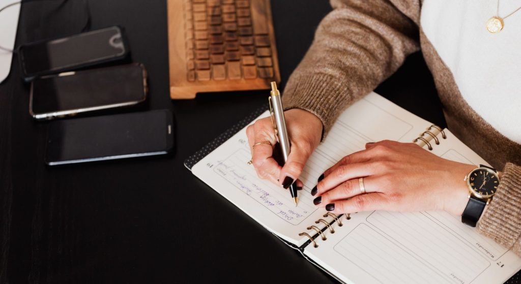 A lady food journaling with a metal pen on a black desk.