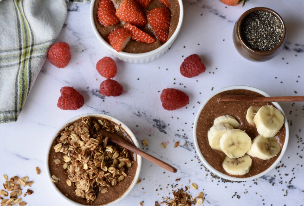 Chocolate Chia Protein Pudding in three white bowls with three different toppings.  One is topped with banana, one with sliced strawberries, and one with homemade granola.