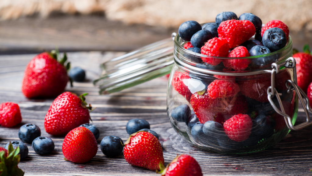 Eat berries for weight control and a fiber rich diet.  Raspberries and blueberries in a glass jar with strawberries beside it on a board.