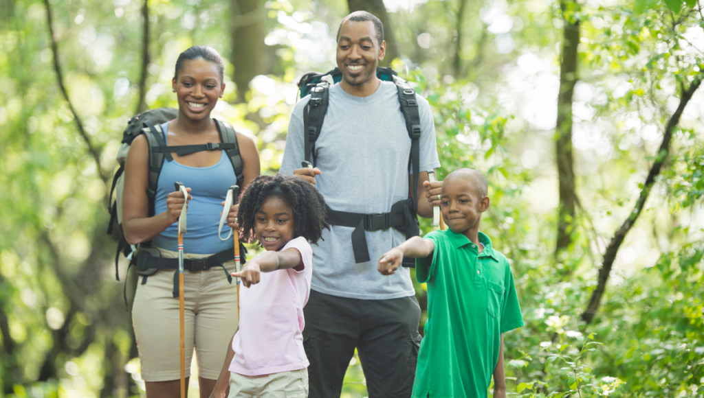 Improve your health and take a vacation. Pictured is a family of four hiking with two kids pointing at something in the woods.