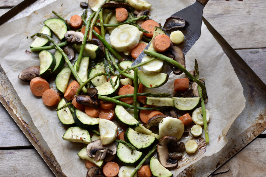 Roasted vegetables and mushrooms side dish scooped up with a metal spatula on brown parchment paper.