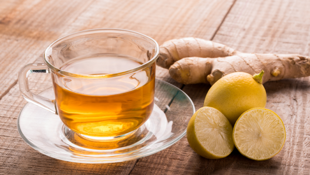 A clear mug of tea with fresh lemon and ginger sitting beside it.
