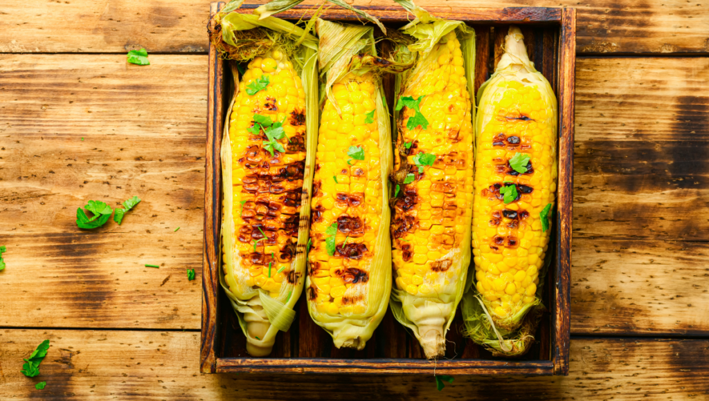 Four grilled corn on the cob in a wooden tray.